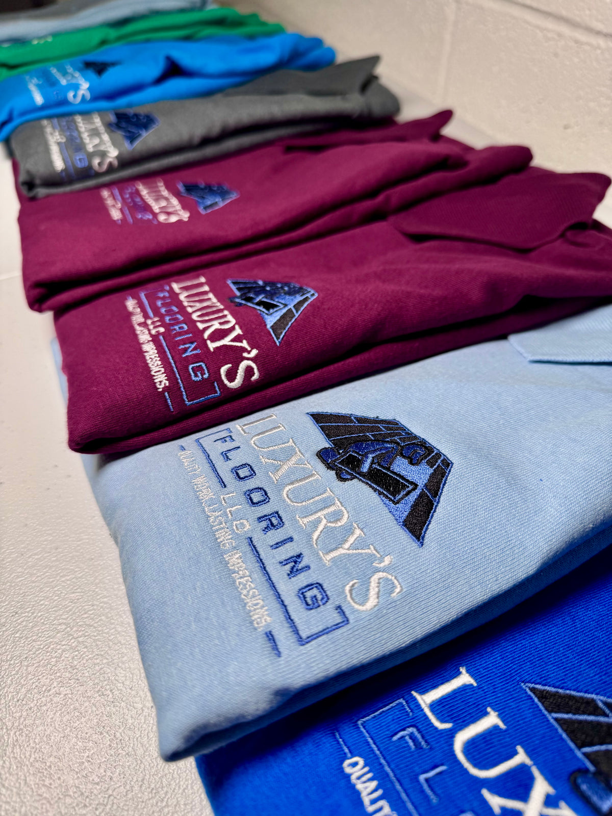 A stack of folded t-shirts in various colors, including blue, maroon, gray, green, and navy, each printed with the logo and text 'LUXURY'S FLOORING' on the chest, arranged diagonally on a light-colored floor.