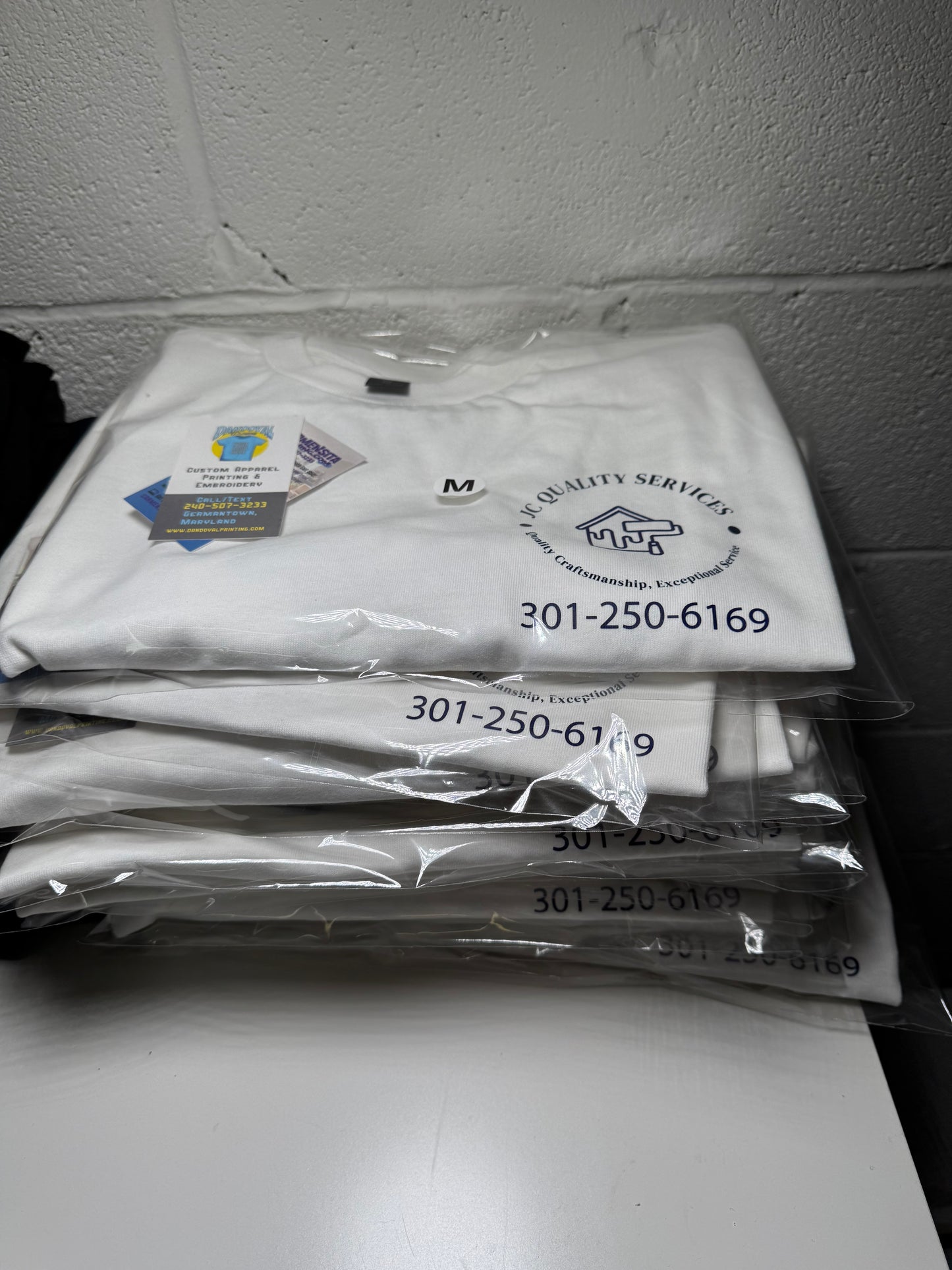 White t-shirts with logos and text on a shelf against a tiled wall.