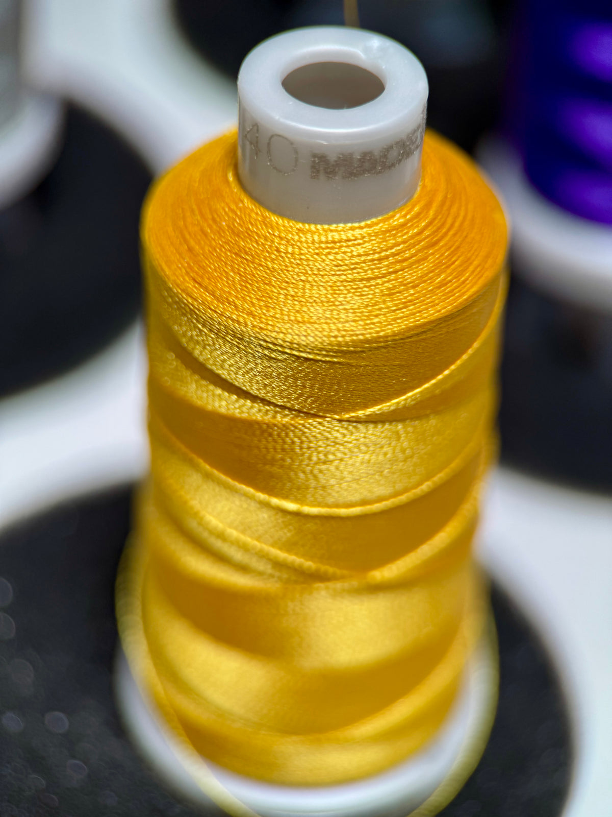 A close-up of a spool of bright yellow satin thread, with the white plastic core showing partial branding text, set against a blurred background of other spools.