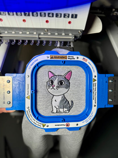 Embroidery machine with a cartoon cat design on a gray fabric