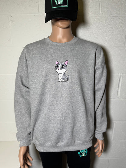 Person wearing a gray sweatshirt with a cat graphic and a black cap against a white wall.