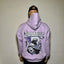 Person wearing a purple hoodie with a graphic design and text, standing against a white brick wall.