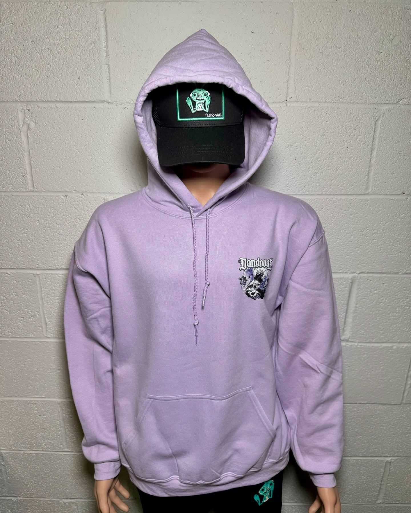 Person wearing a light purple hoodie with a logo, standing against a plain wall.