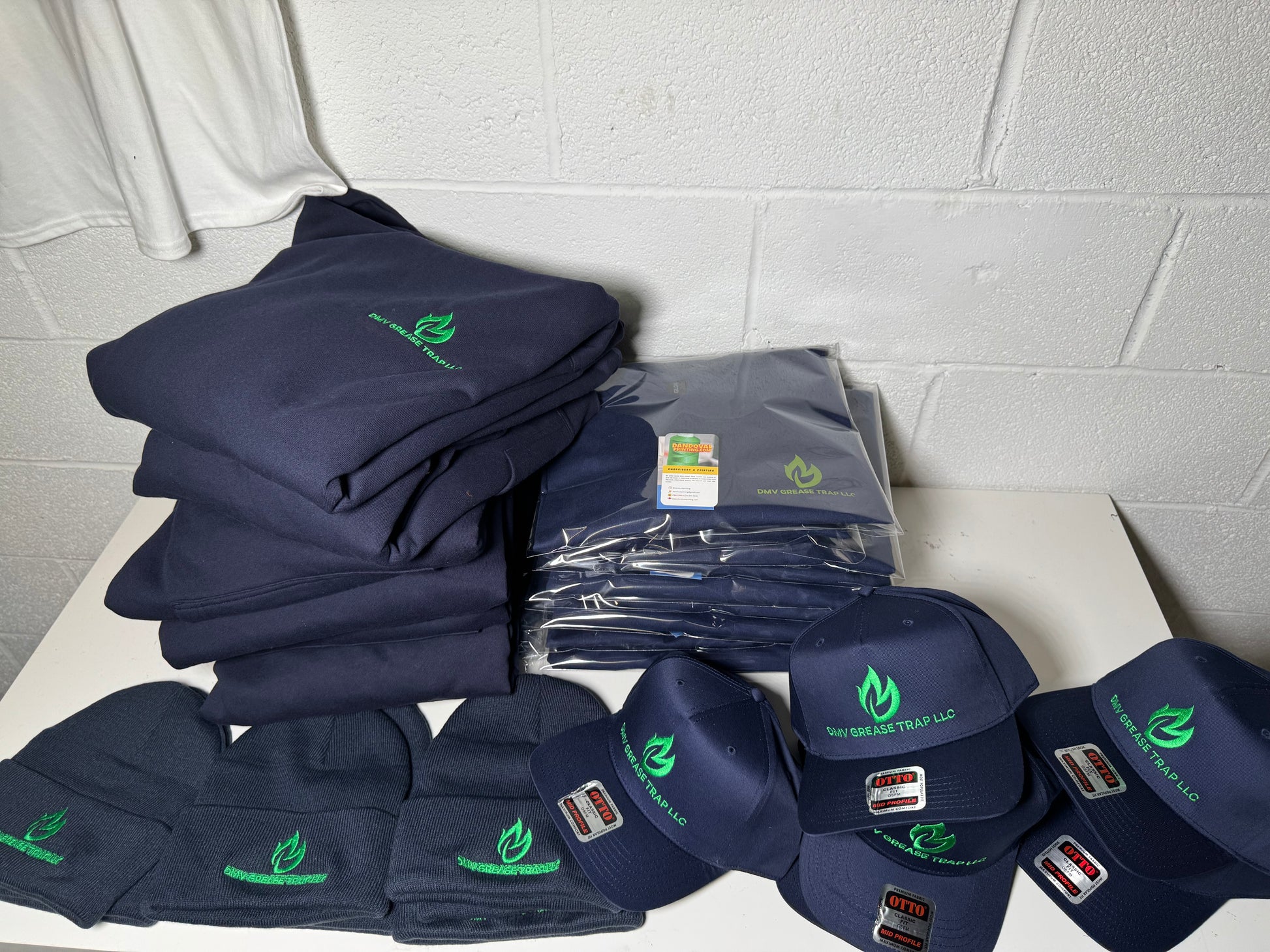 Stack of navy blue shirts, hats, and other items with logos on a white surface against a gray tiled wall.